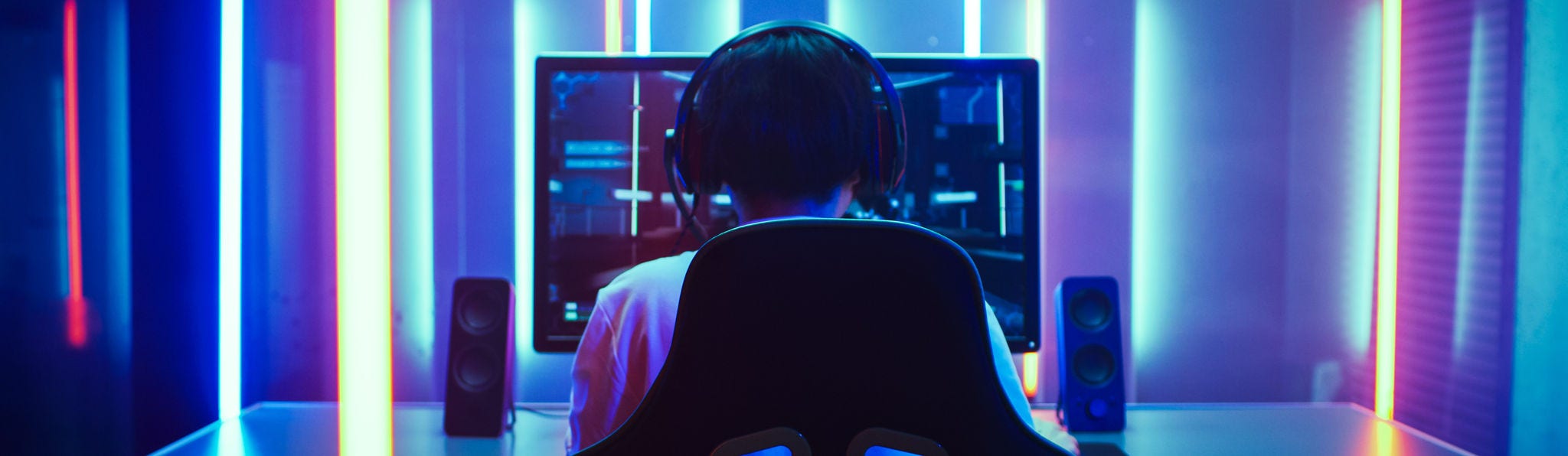 Back View Shot of the Beautiful Professional Gamer Girl Putting on Headset and Starts Playing Online Video Game on Her Personal Computer. Cute Casual Geek Girl. Room Lit by Neon Lamps in Retro Arcade Style.