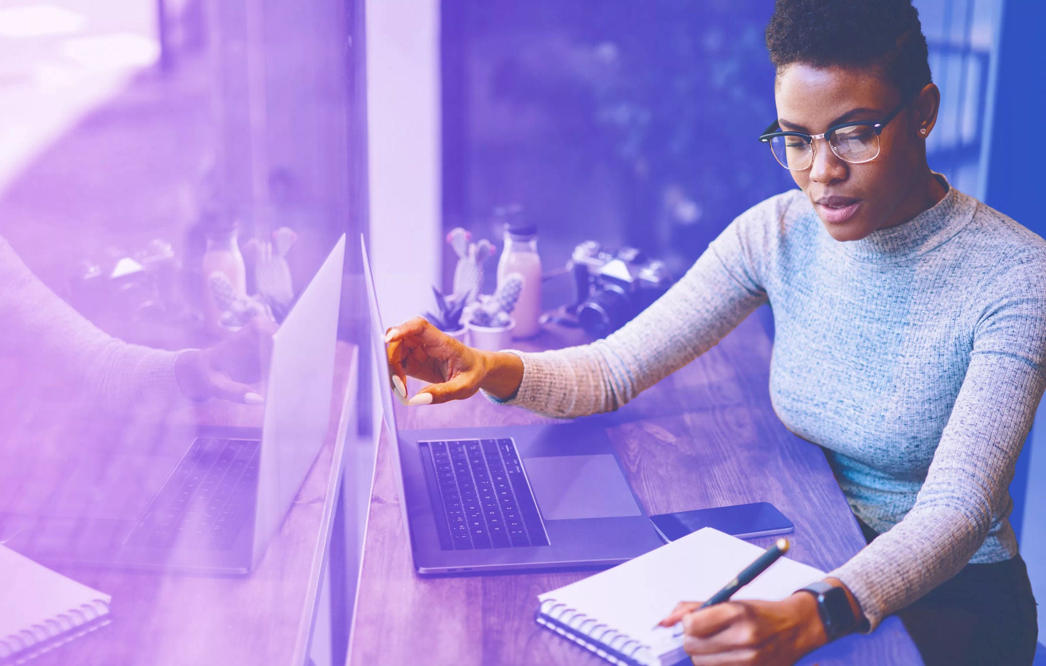 Mulher numa secretária com a mão direita a apontar para o portátil e a outra a tirar notas num bloco