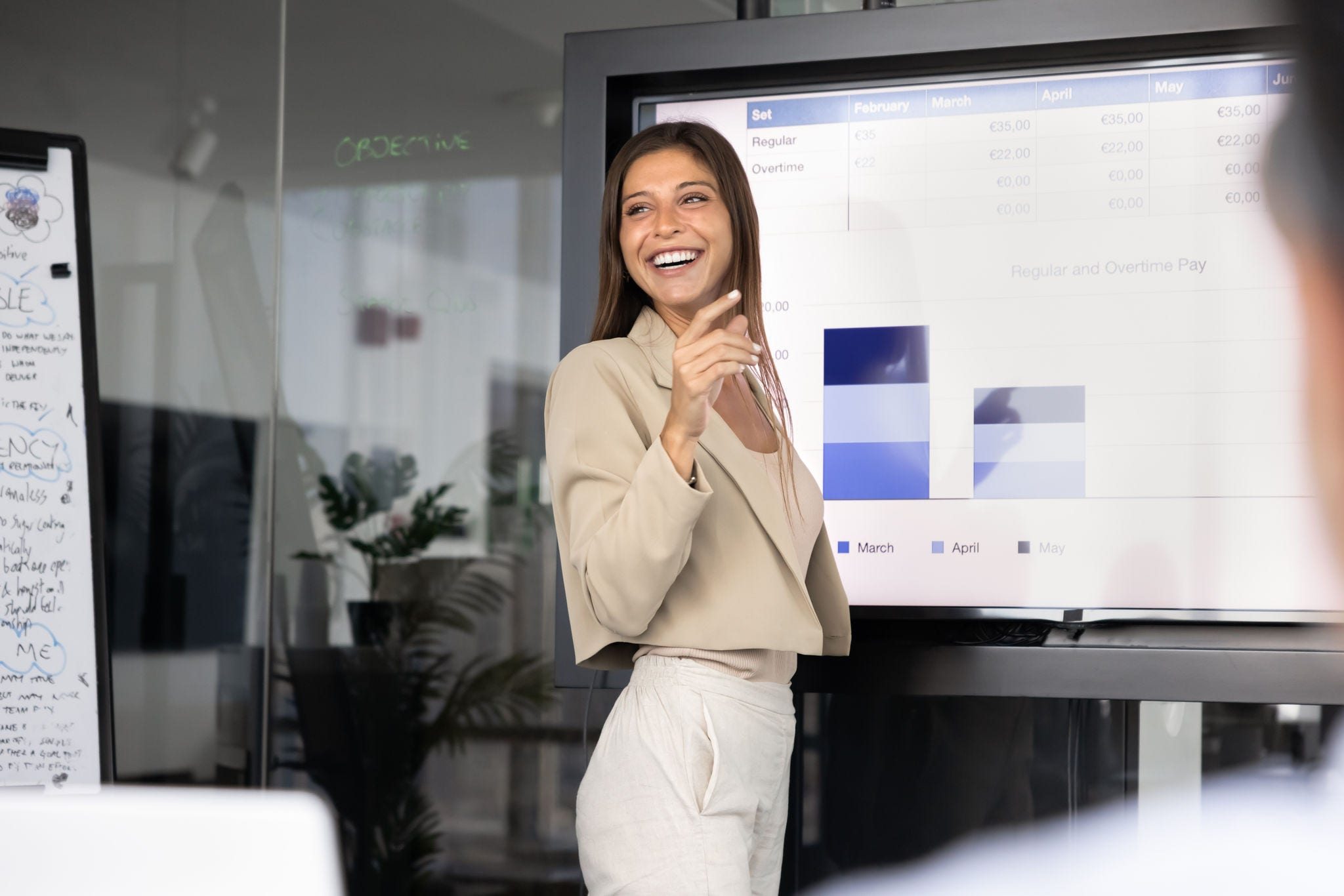 Happy young female business product manager presenting analytic data on whiteboard to team of colleagues, partners, standing at large screen with graphs, smiling, laughing, holding meeting