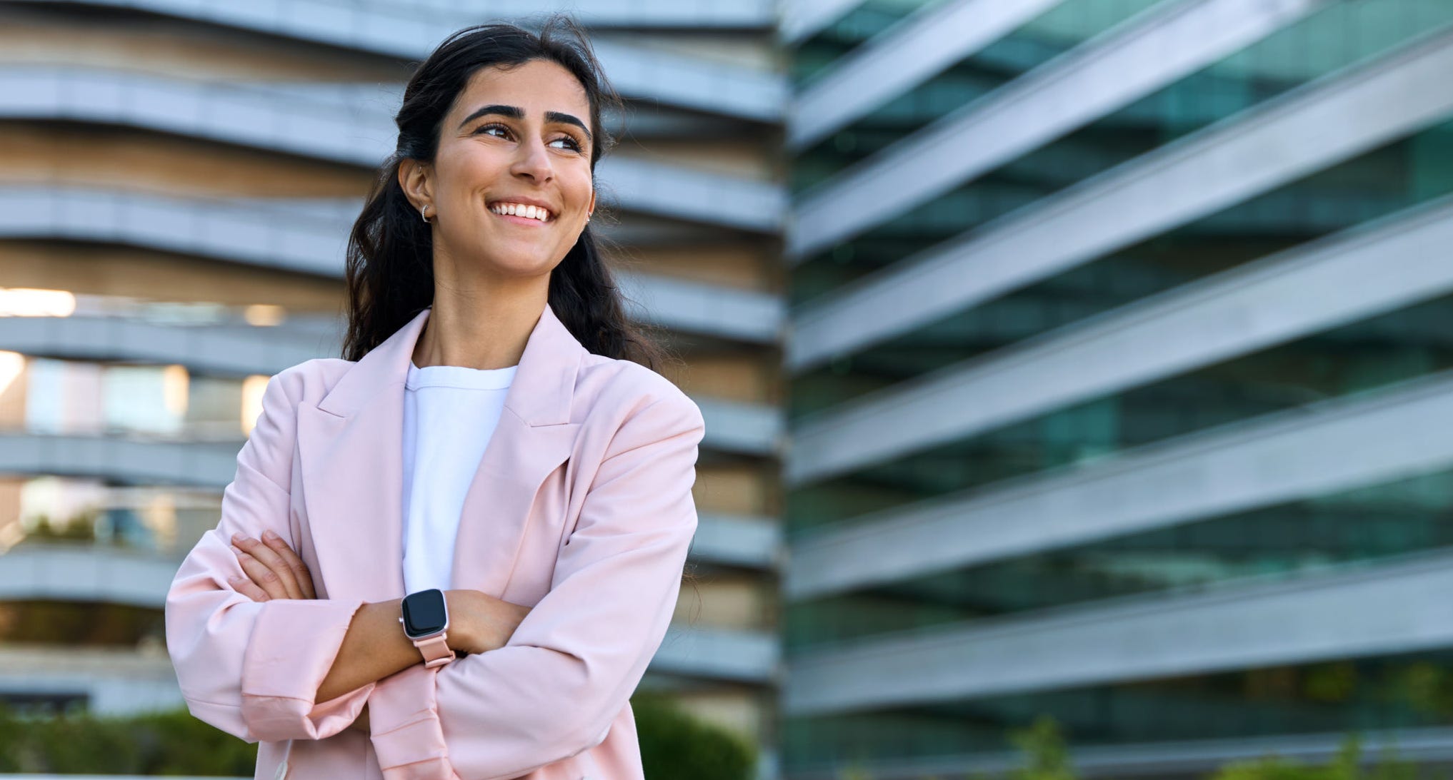Confident portrait of young middle eastern Israel businesswoman standing with crossed hands at office building. Successful smiling indian or arabic woman in business suit looking aside. Copy space