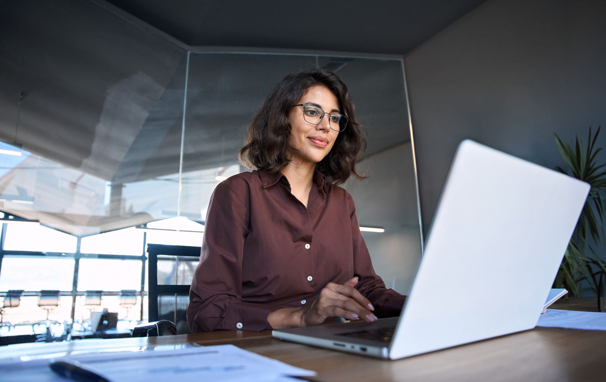 Young professional it specialist latin hispanic business lady working on laptop pc sitting at desk in modern office space. 30s middle eastern indian woman using computer technology app for work online