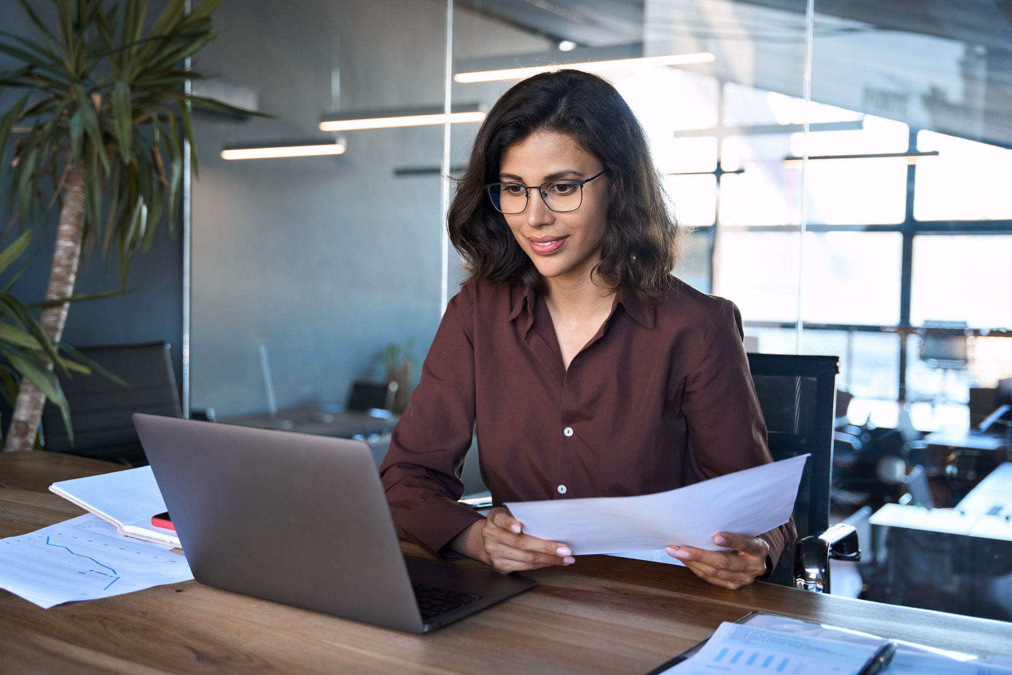 Focused latin hispanic young business woman working on laptop computer reading financial document report in office. Accountant entrepreneur manager businesswoman doing paperwork using pc. Copy space