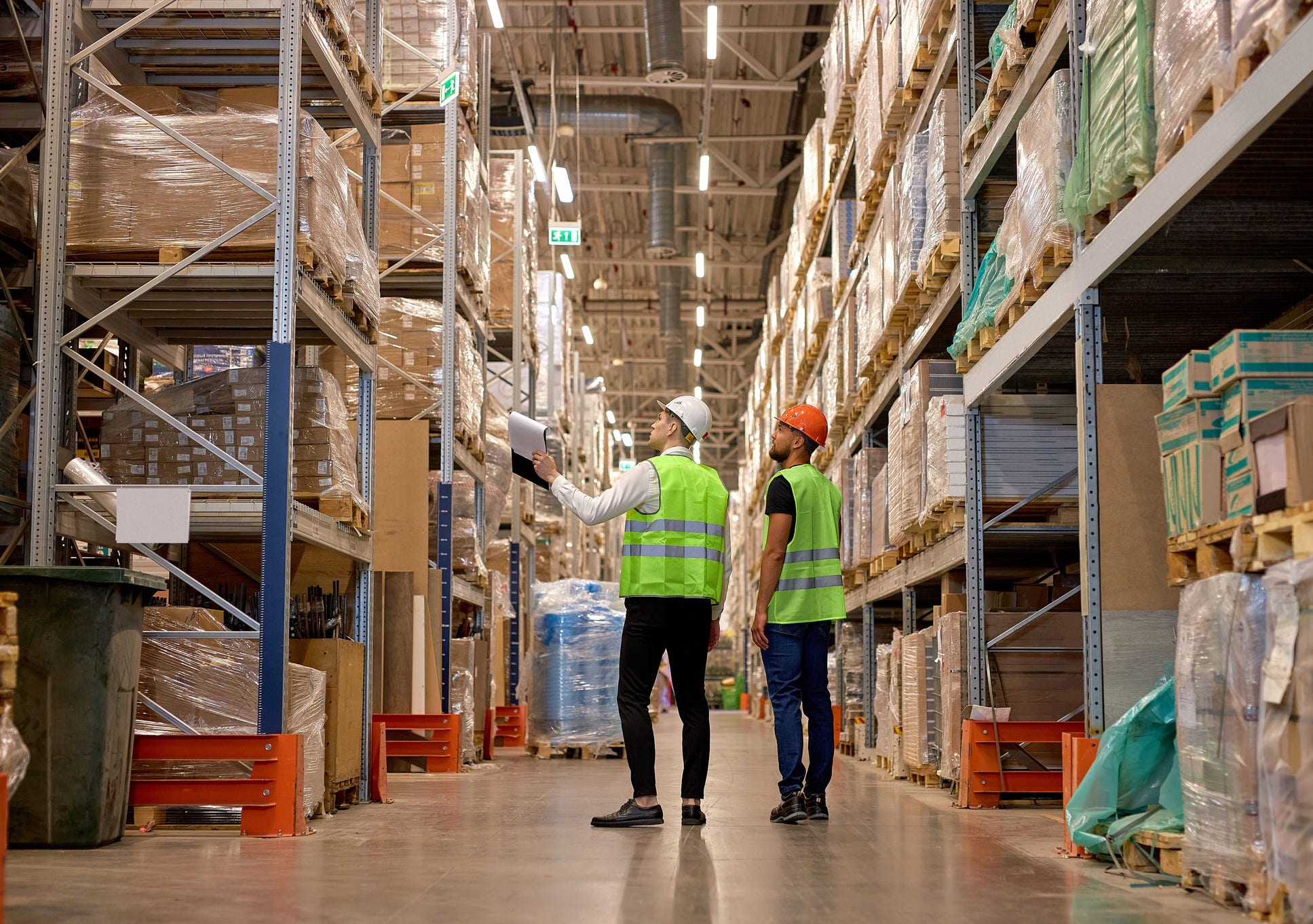two men in warehouse wearing safety helmet, vest. Concept for industry, job, meeting, work training. Two caucasian warehouse workers walking in distribution storage area discussing
