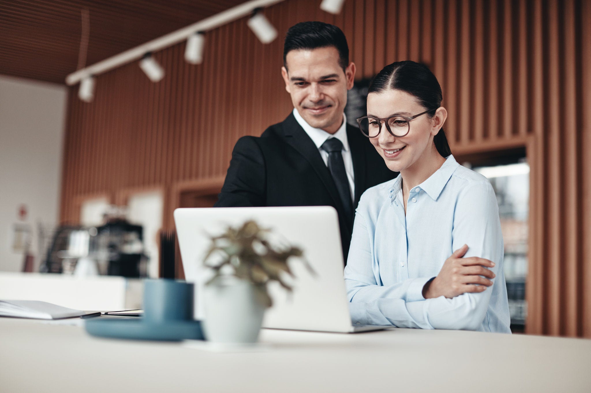 Two young businesspeople smiling while standing at a table going over notes and working on a laptop in an office