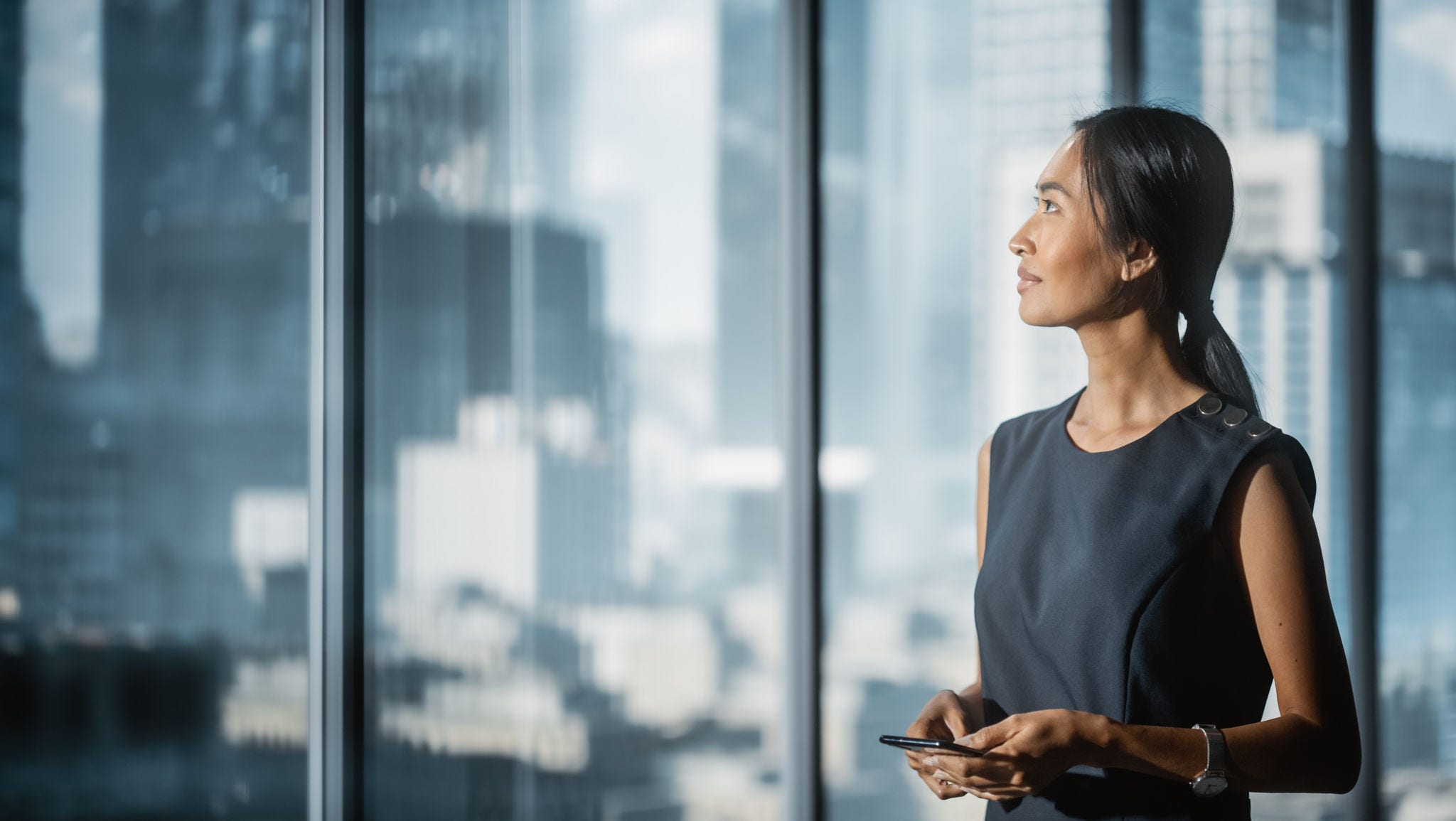 A woman standing beside floor-to-ceiling windows and holding a smartphone, with an urban skyline visible in the background