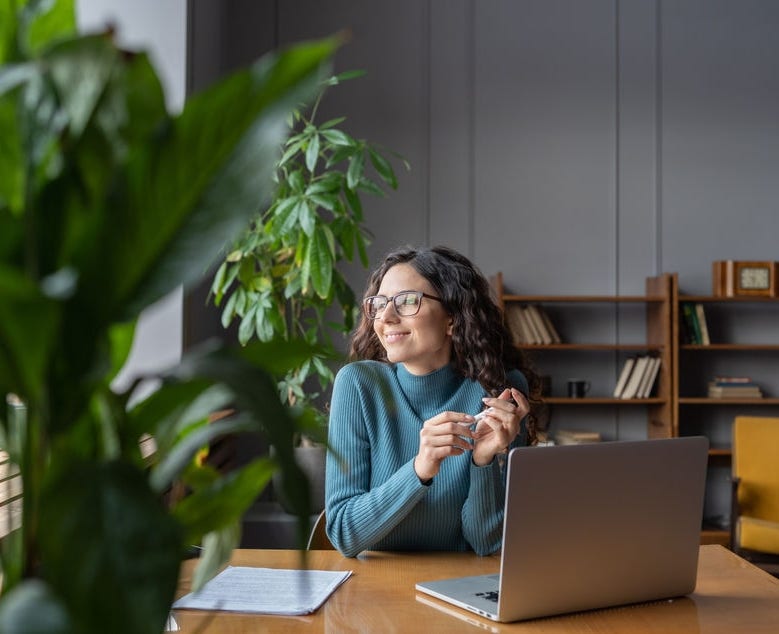 Happy female employee looking in window with satisfied face expression while sitting in office in front of laptop, woman taking break from computer work at workplace. Job satisfaction concept