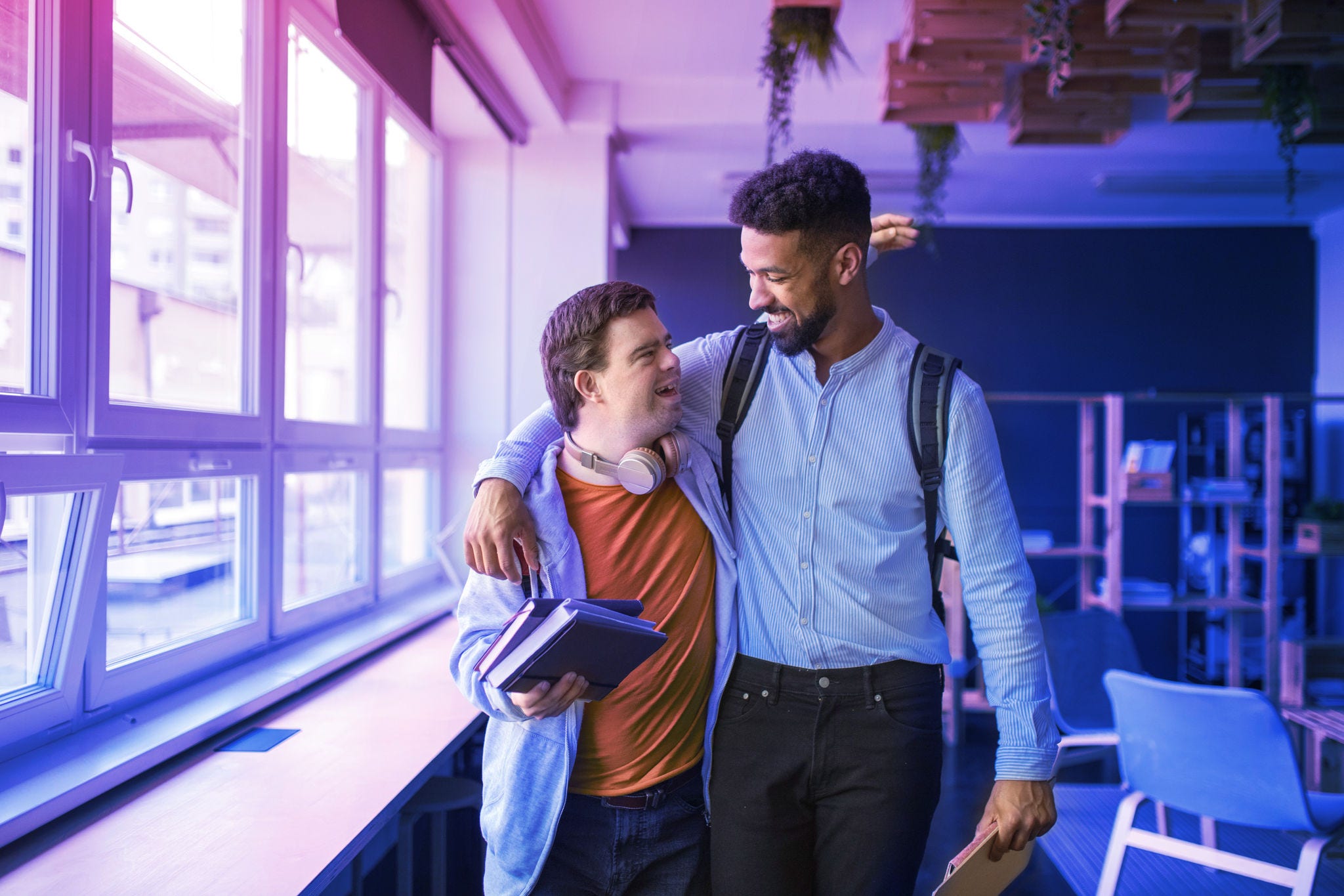 A young man with Down syndrome and his tutor with arms around looking at each other indoors at school