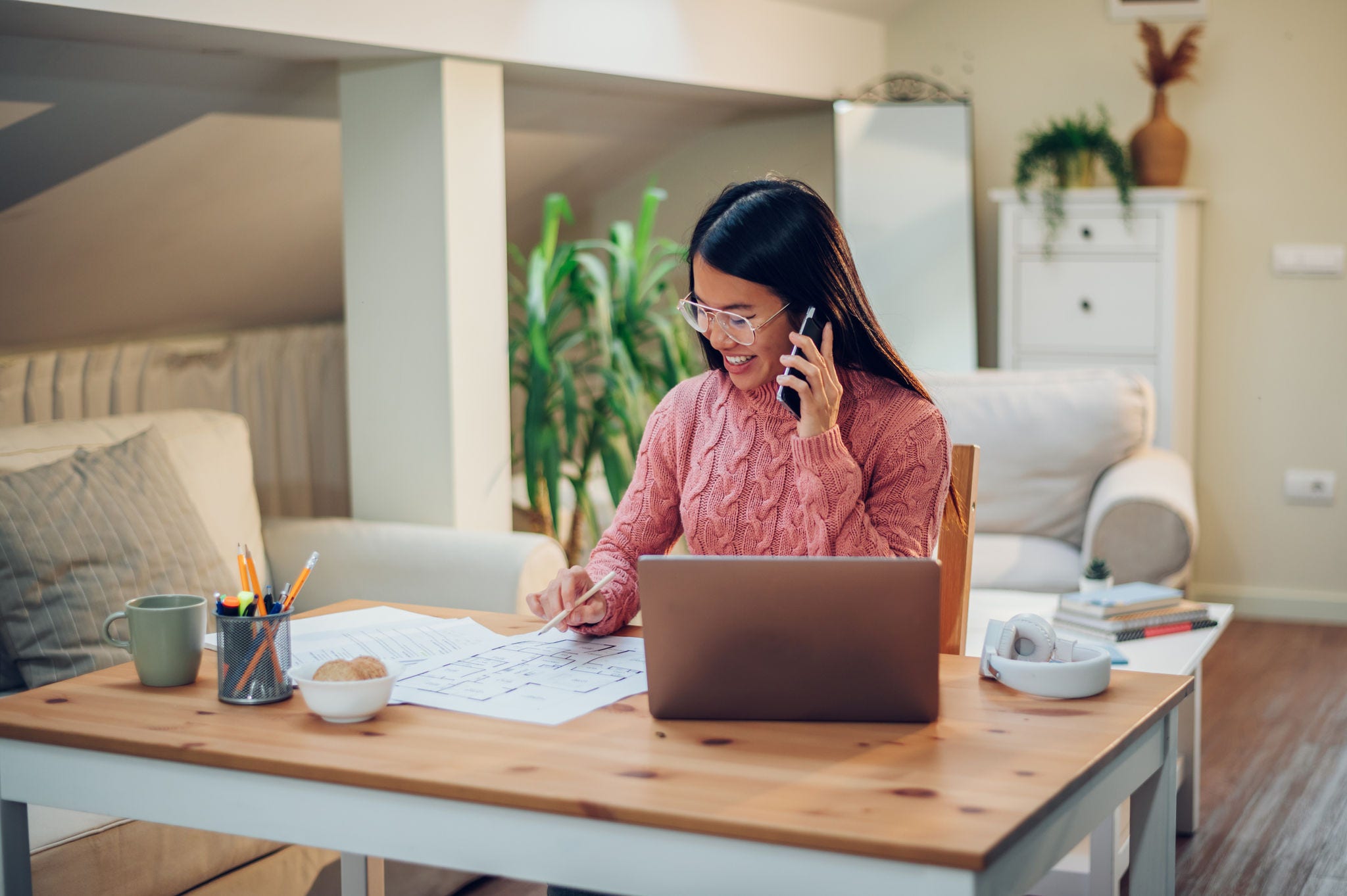 Young Vietnamese woman working in a home office and using a laptop and talking on a smartphone. Flexible workspace and working time. Beautiful Asian business female or student multitasking.