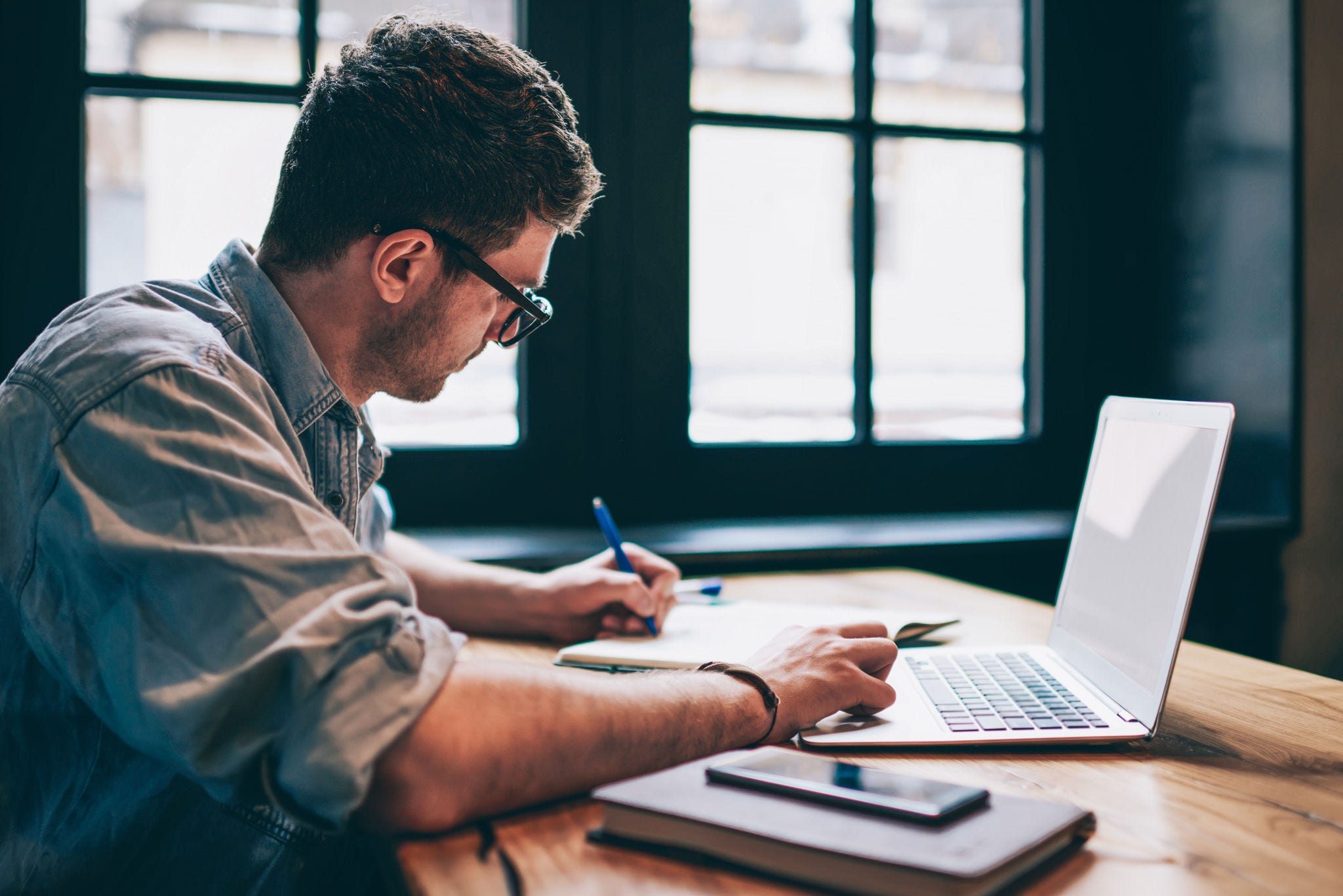 Young millennial male student searching information for homework using laptop and wifi connection indoors, man writing article in textbook for add interesting content on own website or web page