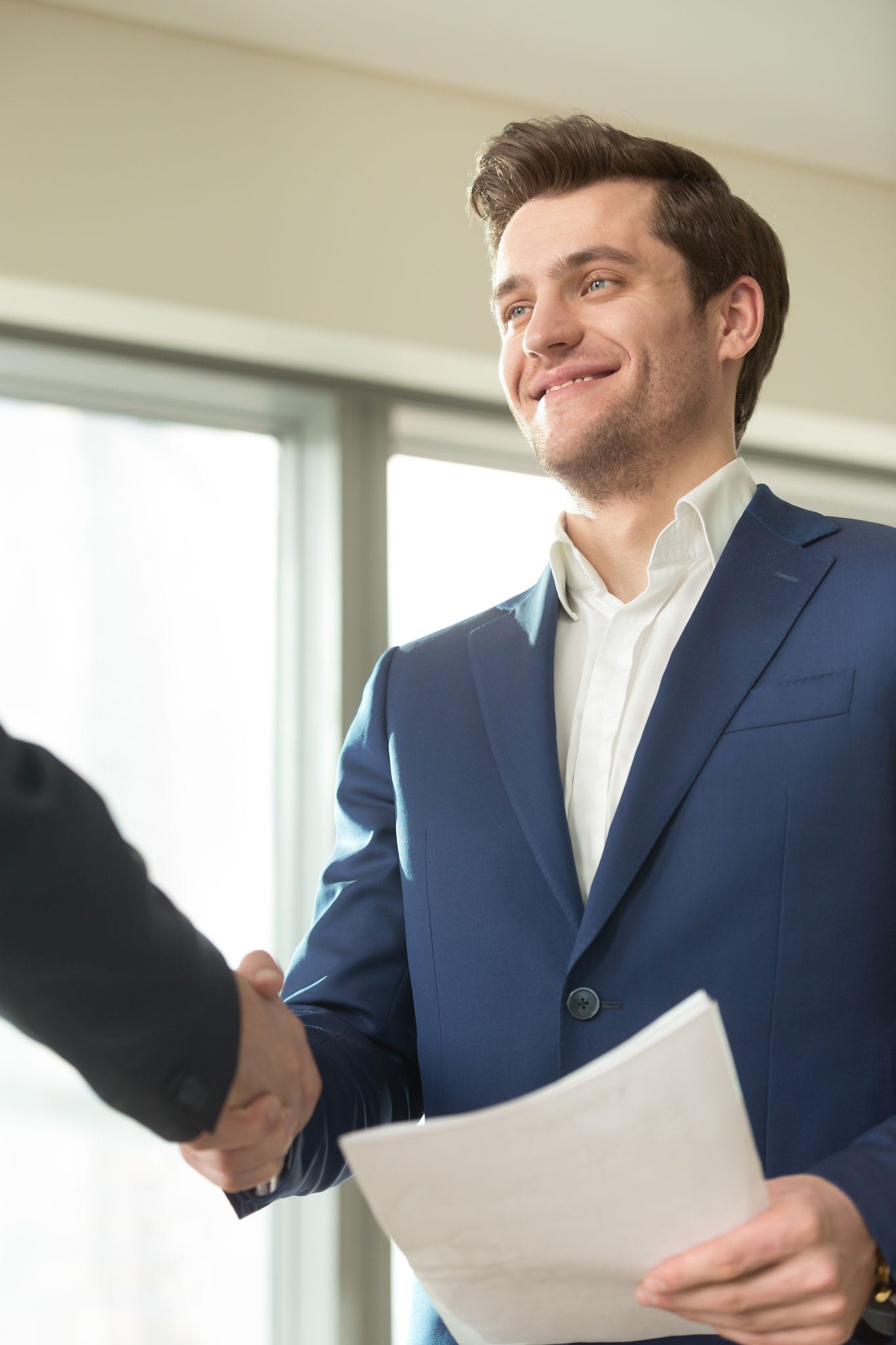 Confident smiling businessman with documents in hand handshaking with partner at business meeting in office. Presentable financial consultant welcoming client, congratulating investor with good deal