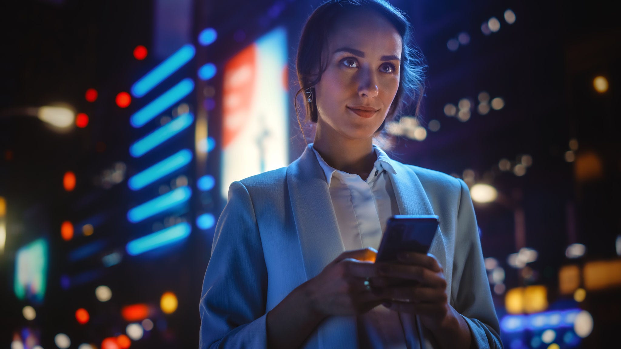 Beautiful Young Woman Using Smartphone Standing on the Night City Street Full of Neon Light. Portrait of Gorgeous Smiling Female Using Mobile Phone.