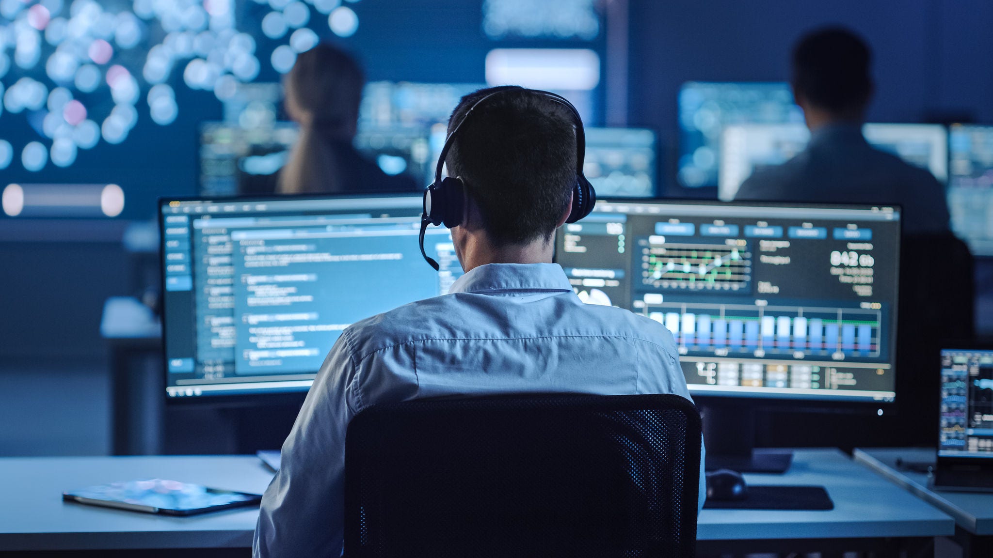 Confident Male Data Scientist Works on Personal Computer in Big Infrastructure Control and Monitoring Room with Neural Network. Young Engineer in an Office Room with Colleagues.