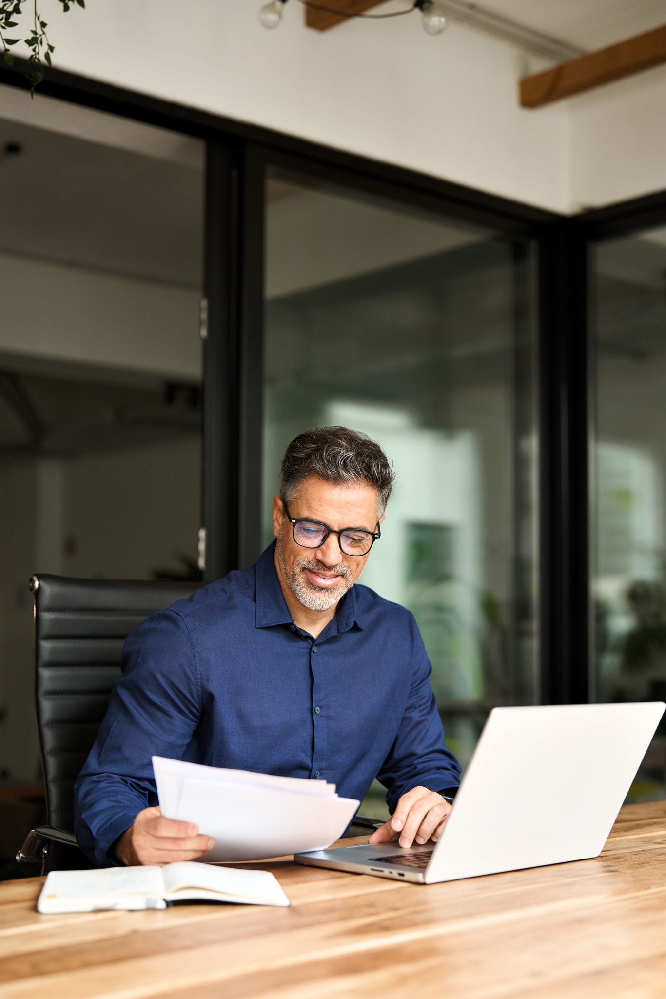 Busy middle aged professional business man using laptop computer reading document at work. Senior mature manager executive or lawyer working on laptop checking paper finance report in office. Vertical