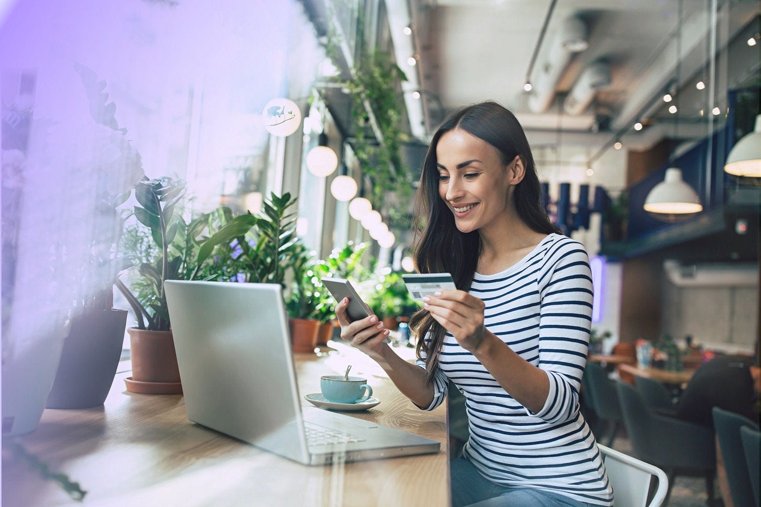 Woman in a café with a computer and mobile phone smiling while holding a credit card