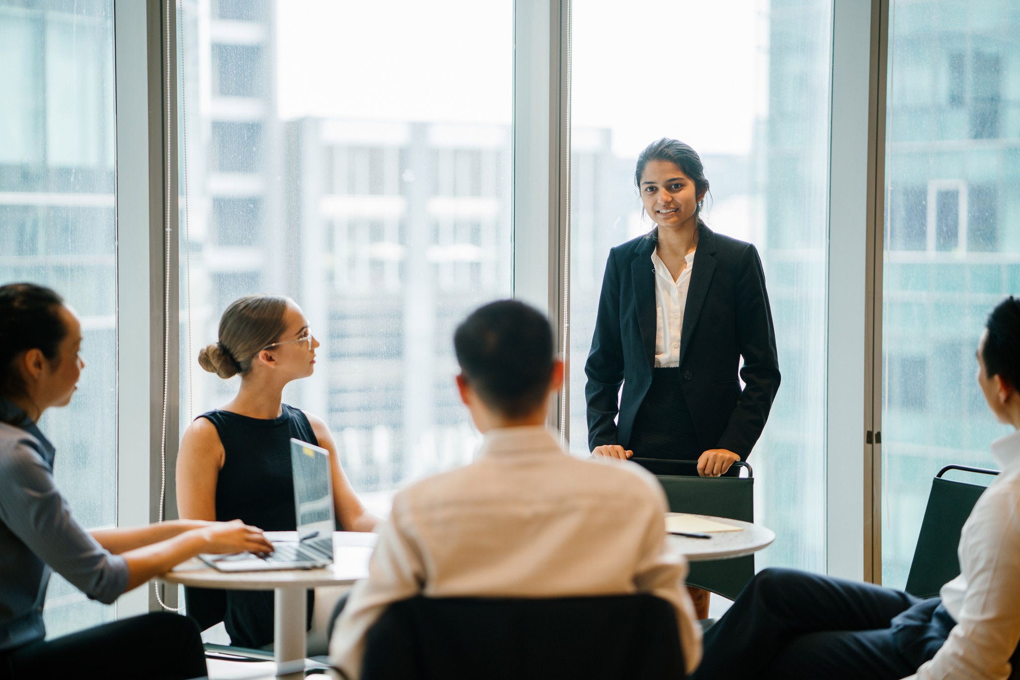 An Asian Indian businesswoman is having a conversation with her colleagues in a conference room and looking forward for their project. She's wearing a black blazer and white blouse with a black skirt.