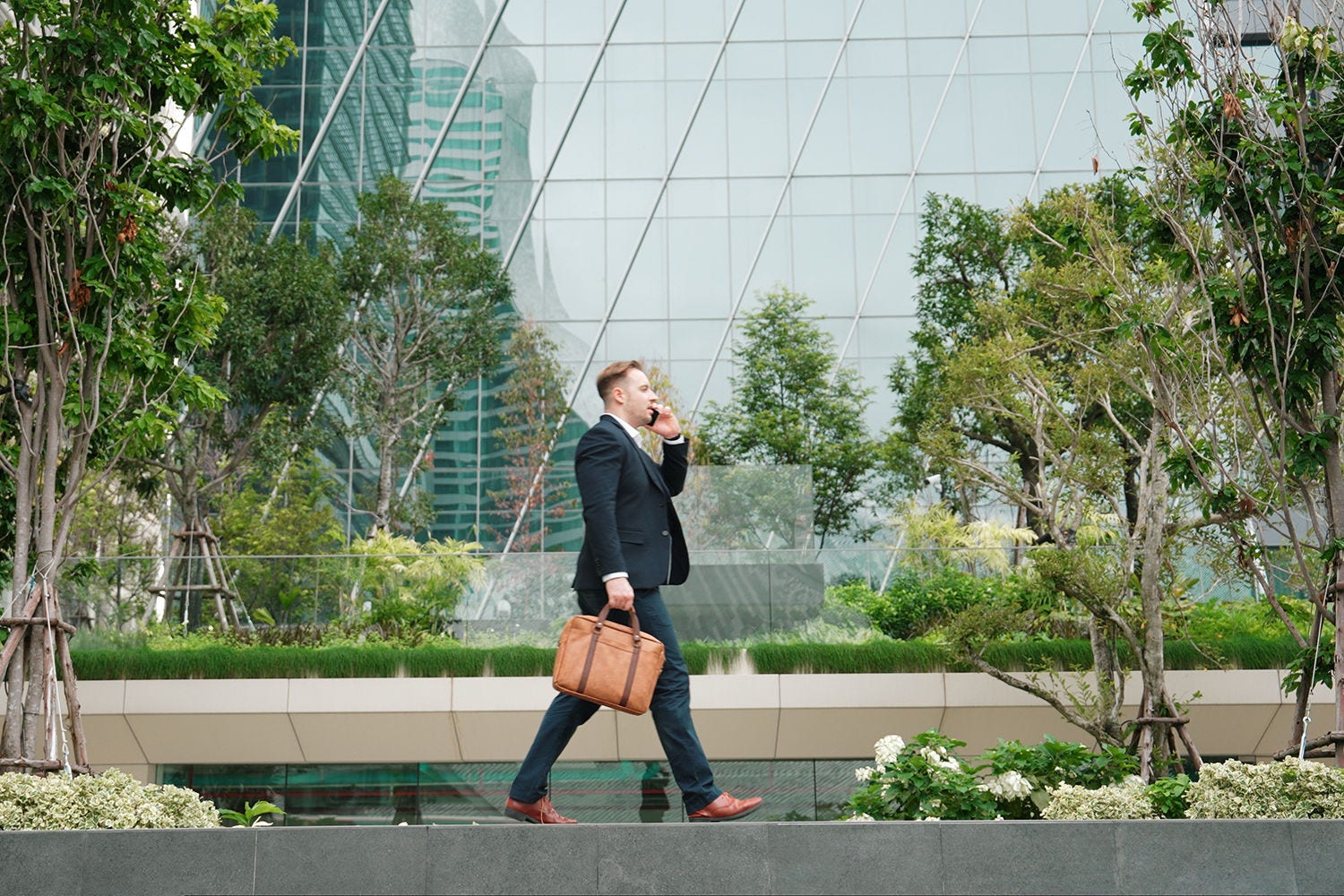 Side view of businessman calling manager about marketing report and walking at modern building in green eco city. Leader going to workplace while talking to his team by using phone. Lifestyle. Urbane.