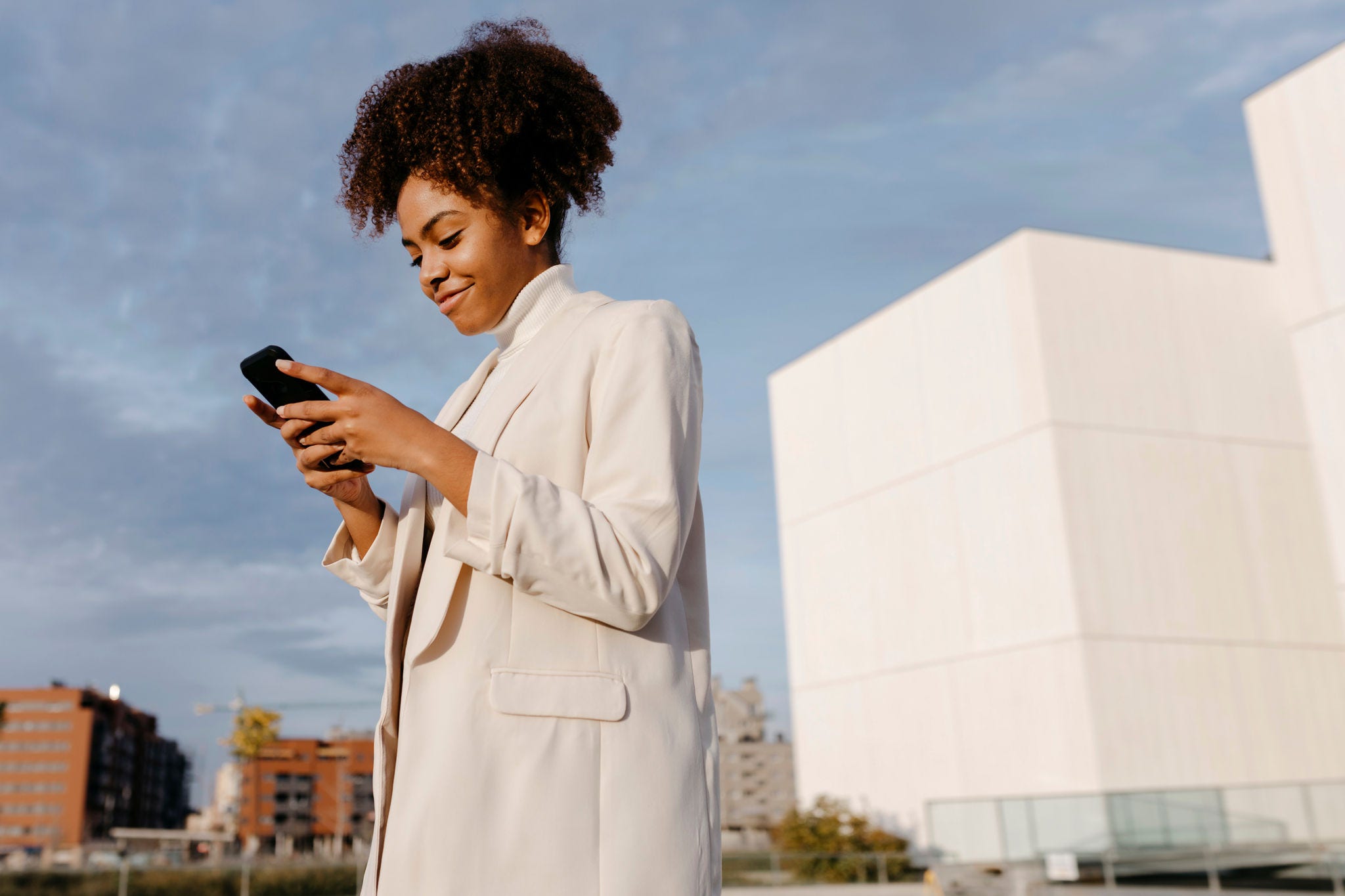 Smiling young woman with afro hair using smart phone while standing against sky in city
