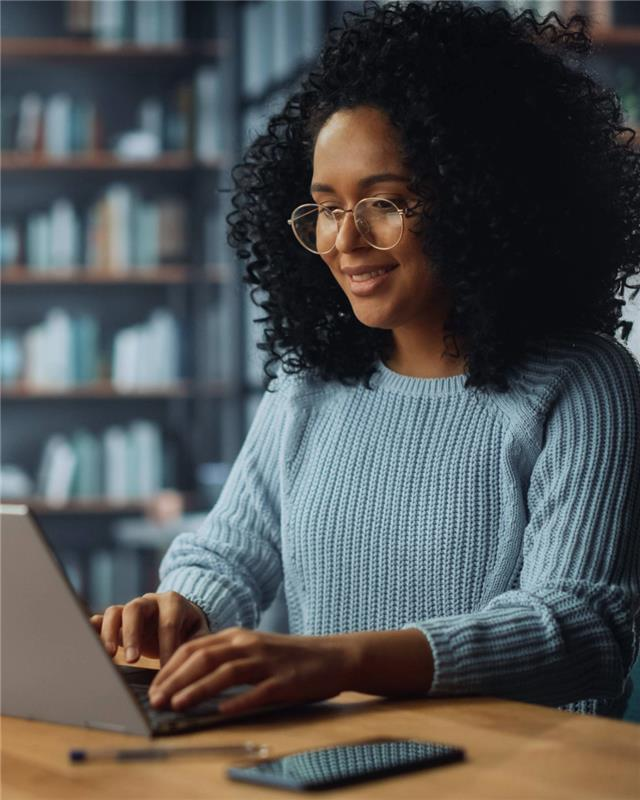 A person wearing glasses typing on a laptop in a library