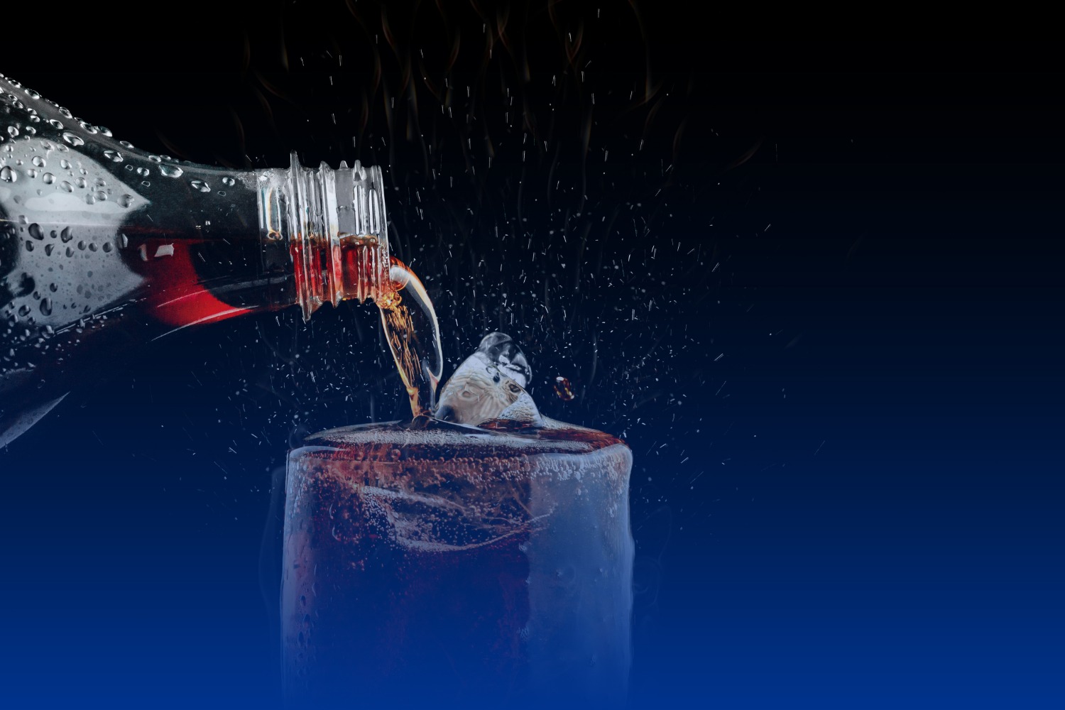 Bottle pouring a dark fizzy drink into a glass with ice, with droplets and splashes against a dark background.